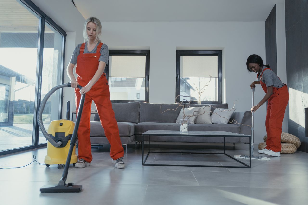 hero-img-02 Two women in red overalls vacuuming and sweeping a modern living room.